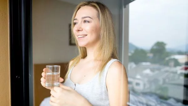 mulher feliz olhando para janela com água retirada do purificador em copo de vidro